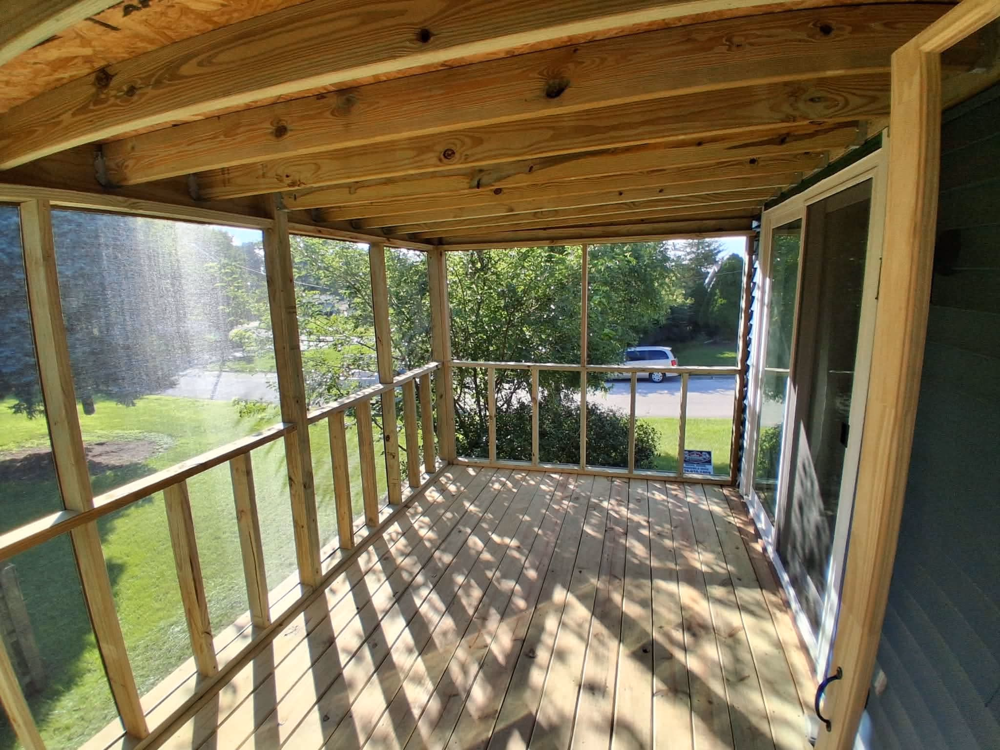 Empty wooden screened porch with sunlight and shadows, overlooking a green yard and a parked car.