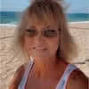 Woman with sunglasses smiling on a sandy beach with the ocean in the background.